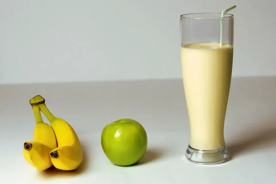 A banana shake in a tall glass with a banana and an apple