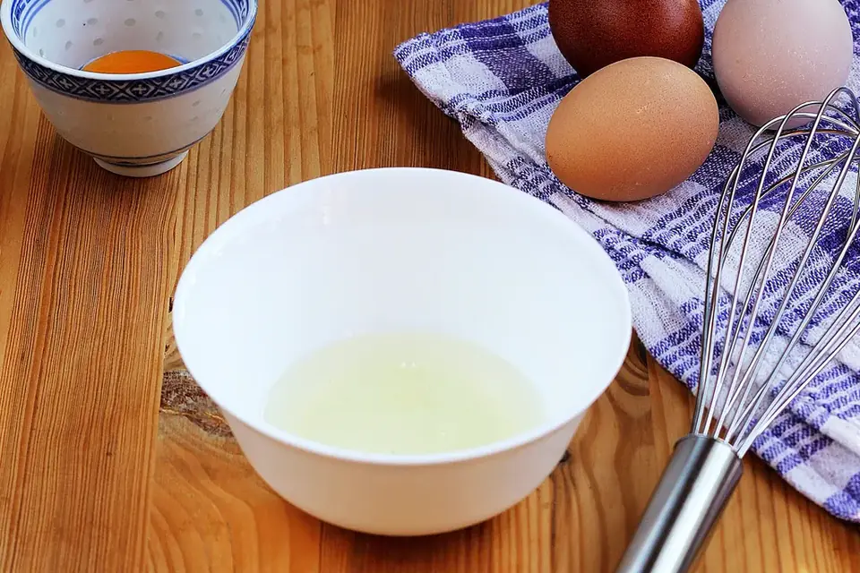 Separated egg whites in a bowl with eggs and a whisk